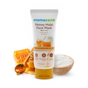 Mamaearth Honey Malai Face Wash tube (100 ml) standing upright against a white background, surrounded by honeycomb, a wooden spoon dripping with honey, and a bowl of creamy malai (milk cream). The label highlights key ingredients—Honey and Malai—and mentions it is Made Safe certified, dermatologically tested, and suitable for all skin types. Tagline “For Nourishing Glow” is prominently displayed.