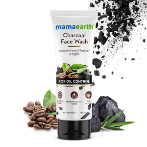 Mamaearth Charcoal Face Wash tube (100 ml) placed against a white background with visuals of coffee beans, green leaves, charcoal chunks, and black charcoal dust particles. The label highlights key ingredients—Activated Charcoal and Coffee—and emphasizes oil control benefits. Product features include "Made Safe Certified," "Dermatologically Tested," and "For Oily Skin."